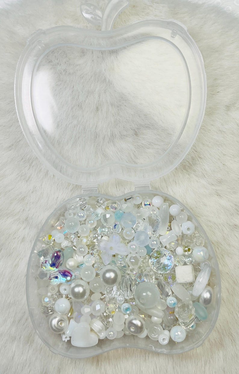May include: Clear, apple-shaped container filled with an assortment of white and iridescent beads, pearls, and decorative elements. The beads vary in size and shape, including round, faceted, and floral designs. The container is open, revealing the contents.