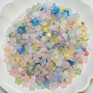 May include: A collection of pastel-colored flower and star-shaped beads in shades of pink, blue, yellow, and green. The beads have a translucent quality and are scattered on a white, scalloped-edge plate. Some beads have gold flecks.