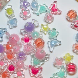 May include: A collection of colorful, translucent beads in various shapes, including flowers, butterflies, and spheres. The beads are in shades of pink, orange, yellow, blue, and purple. Some beads have a swirled or layered design, while others are solid.
