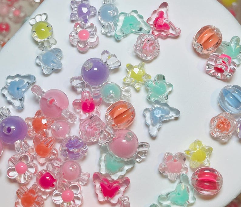 May include: A collection of colorful, translucent beads in various shapes, including flowers, butterflies, and spheres. The beads are in shades of pink, orange, yellow, blue, and purple. Some beads have a swirled or layered design, while others are solid.