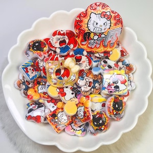 May include: A selection of colourful resin charms in a cartoon style, depicting a well-known cat character in different poses and expressions. The charms are arranged in a white, scalloped-edge dish. Some charms show the character with flames and a computer.