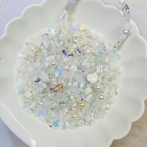 May include: A white scalloped dish filled with an assortment of white, clear, and iridescent beads, including pearls, butterflies, and various shapes. A beaded handle arches over the dish, creating a decorative display.