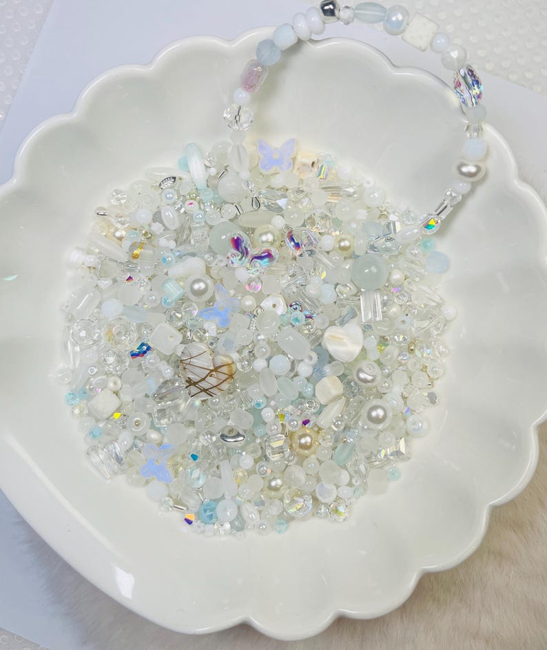 May include: A white scalloped dish filled with an assortment of white, clear, and iridescent beads, including pearls, butterflies, and various shapes. A beaded handle arches over the dish, creating a decorative display.