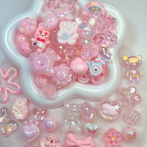 May include: A collection of assorted pink and iridescent beads and charms in various shapes including bears, hearts, bows, flowers, and rabbits. Many pieces feature a glossy finish, and one heart-shaped charm displays the text miss you.