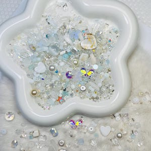 May include: A white, star-shaped ceramic dish filled with an assortment of white, clear, and iridescent beads, pearls, and decorative elements. The mix includes heart-shaped beads, butterfly accents, and various sizes of round and faceted beads. Some beads are scattered around the dish.