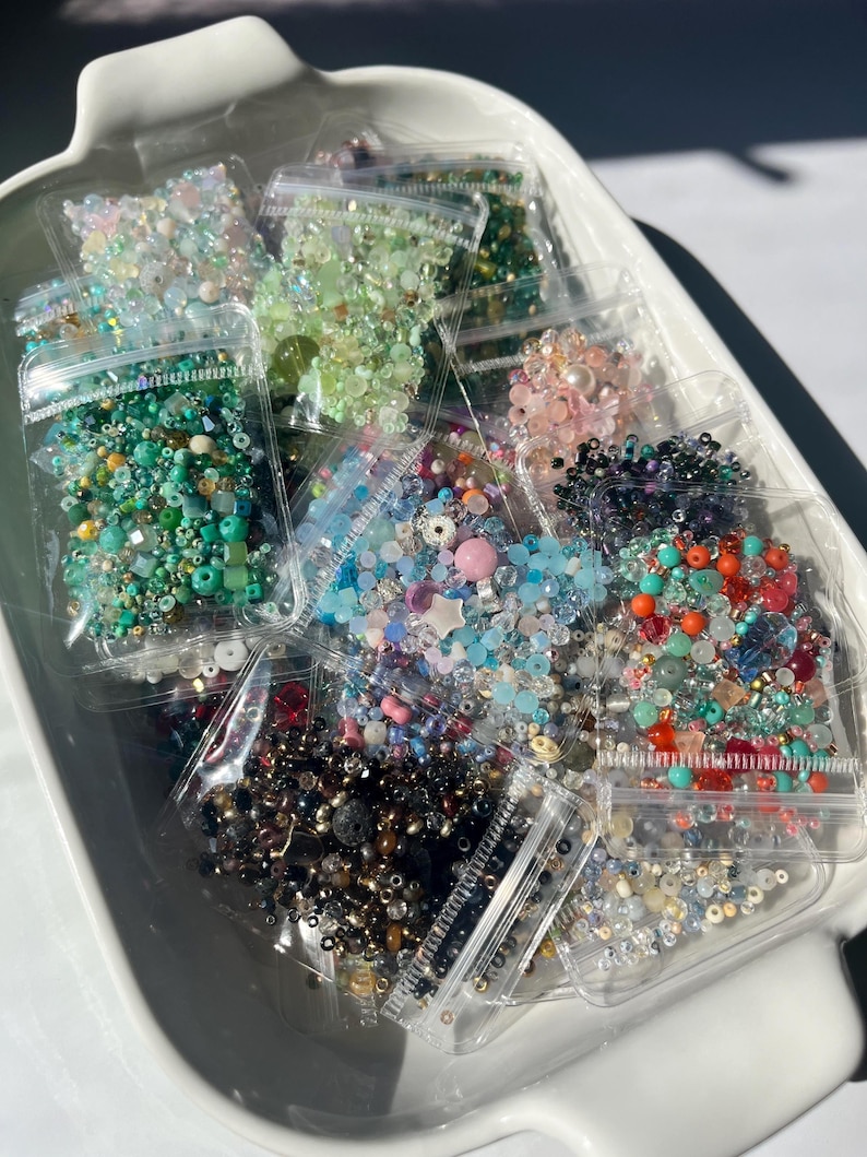 May include: A collection of small, colorful beads in clear plastic bags. The beads are a variety of shapes and sizes, including round, square, and star-shaped. The beads are arranged in a white bowl.