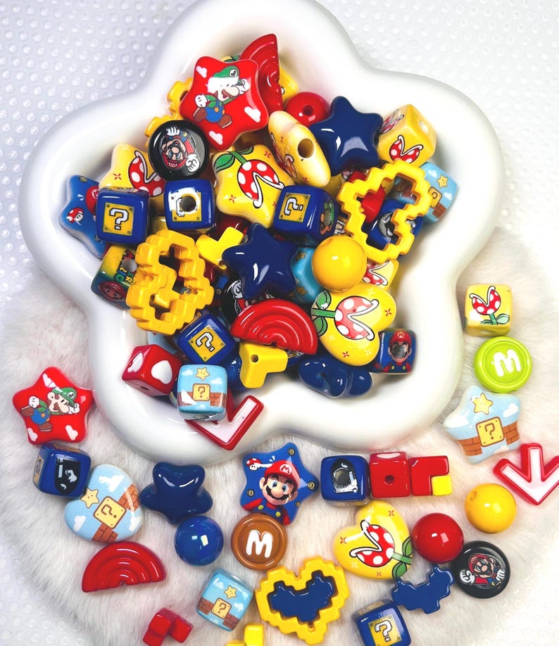 May include: A collection of colorful, themed beads in a white, star-shaped bowl. The beads feature various shapes and designs, including stars, cubes, and hearts, with images of a popular video game character and related icons. The color palette includes red, yellow, blue, and green.