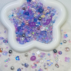 Peut inclure: Un plat blanc en forme d'étoile rempli d'un assortiment de perles violettes, bleues et irisées. Les perles sont de tailles et de formes variées, comprenant des perles, des cristaux facettés et de petits éléments décoratifs. Quelques perles sont éparpillées autour du plat.