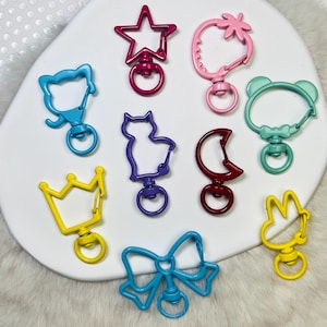 May include: A collection of colorful metal swivel clasps in various shapes, including a star, strawberry, bear, cat, moon, crown, and bow. The clasps are in shades of pink, red, blue, purple, and yellow, with a shiny finish. These clasps are ideal for crafting and jewelry making.