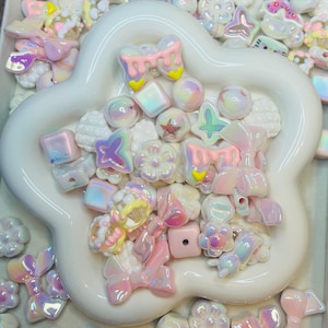 May include: A collection of assorted iridescent and pastel-colored plastic beads in shapes including bows, flowers, stars, and cubes, contained within a white, five-petaled flower-shaped dish, with additional loose beads scattered around the base.