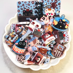 Op de afbeelding: Een verzameling kleine, kleurrijke acrylbedels en een miniatuur spiraalnotitieboekje met cartoon-dieren, waaronder zwarte katten, beren en honden. De items liggen in een witte, geschulpte kom en tonen diverse patronen zoals luipaardprint en teksten zoals TUZI en Keep Lucky.