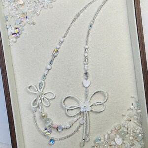 May include: A delicate white beaded necklace with floral and bow designs. The necklace features various sizes of white beads, clear crystals, and a heart-shaped pendant. The necklace is displayed in a wooden-framed tray with scattered beads.