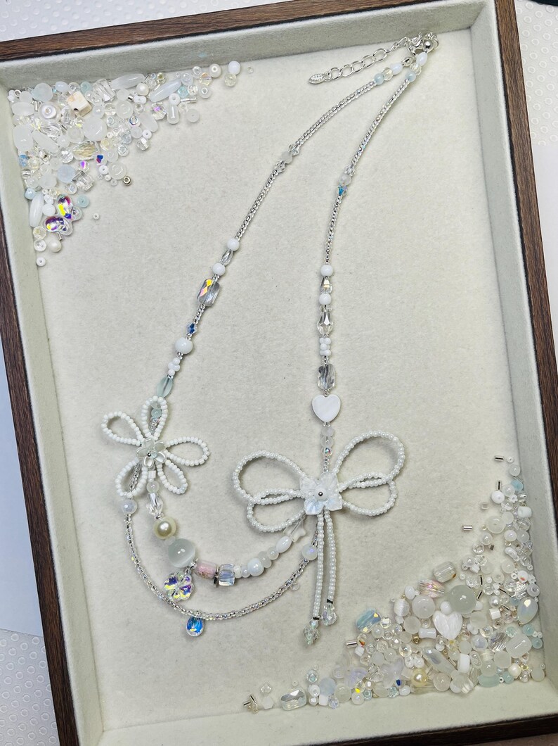 May include: A delicate white beaded necklace with floral and bow designs. The necklace features various sizes of white beads, clear crystals, and a heart-shaped pendant. The necklace is displayed in a wooden-framed tray with scattered beads.
