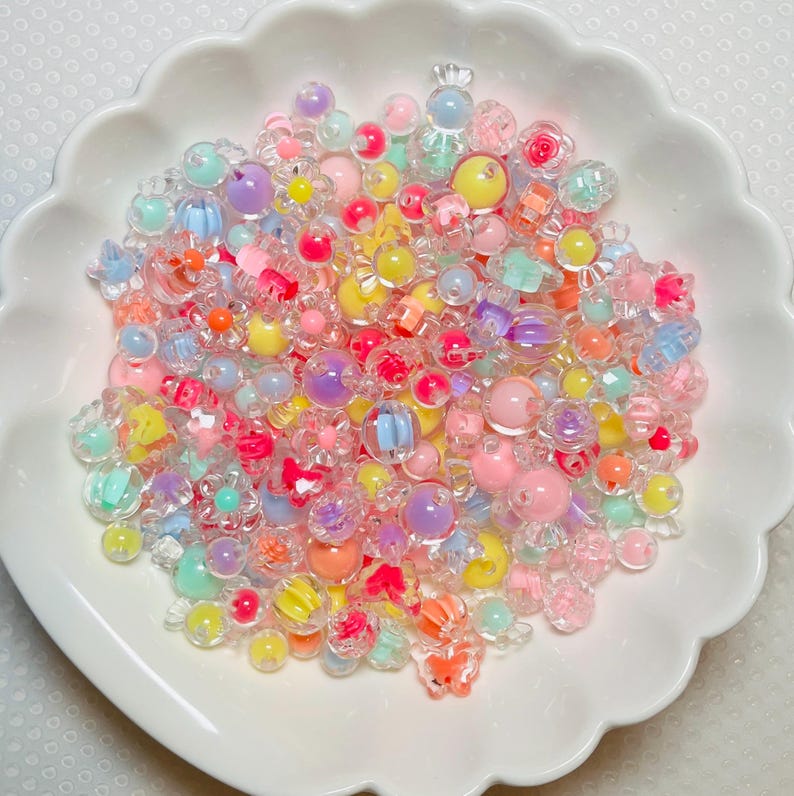 May include: A collection of assorted plastic beads in a white scalloped bowl. The beads feature various shapes including spheres, flowers, and candy forms in translucent and opaque finishes with a vibrant palette of pink, yellow, blue, and purple.