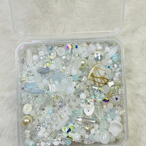 May include: A clear plastic container filled with an assortment of white, iridescent, and clear beads. The beads vary in shape and size, including pearls, crystals, and elongated shapes. A decorative heart-shaped bead with gold accents is also present.
