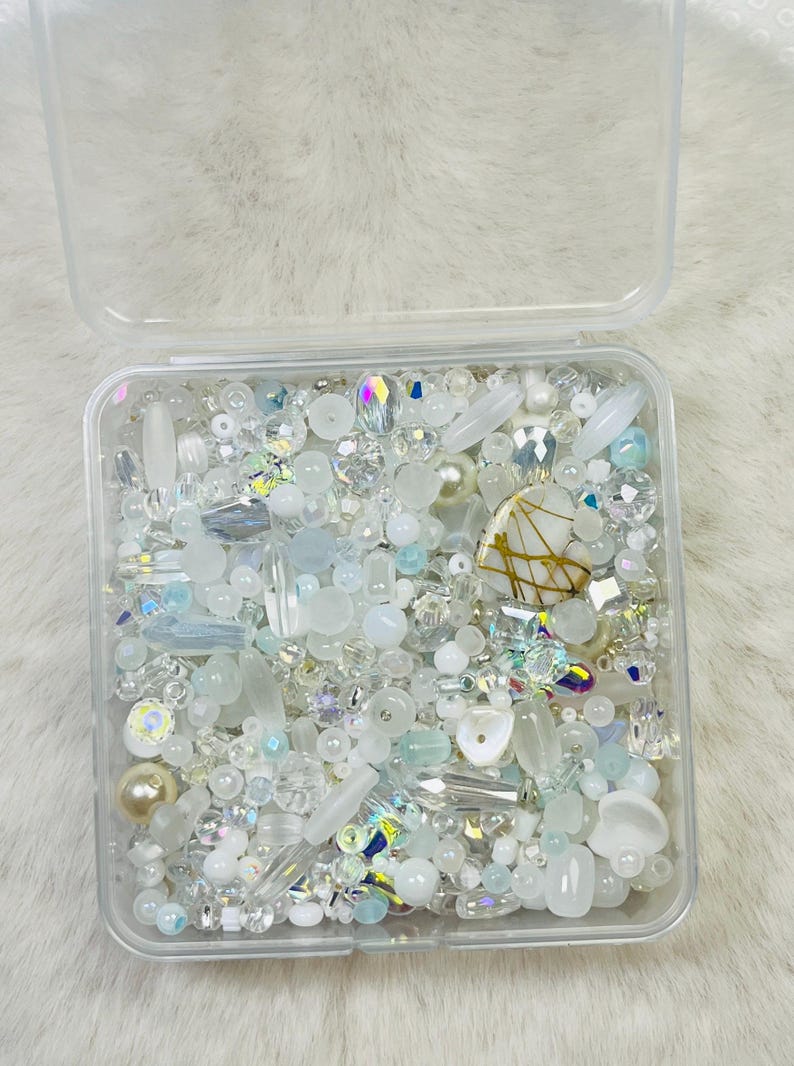 May include: A clear plastic container filled with an assortment of white, iridescent, and clear beads. The beads vary in shape and size, including pearls, crystals, and elongated shapes. A decorative heart-shaped bead with gold accents is also present.