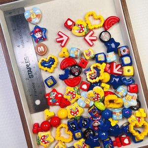 May include: A collection of colorful, themed beads and charms, including stars, hearts, and cubes, with various designs and characters. The assortment features red, yellow, blue, and white elements, with a ruler for scale. The beads and charms are inside a wooden tray.