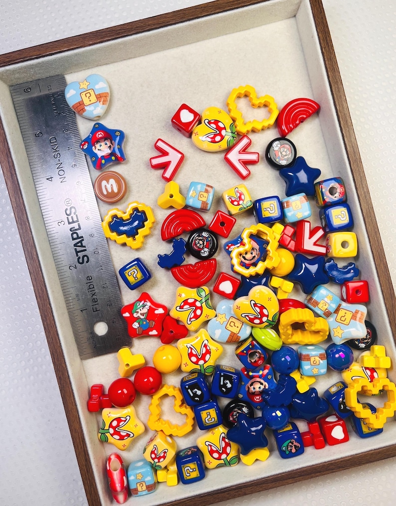 May include: A collection of colorful, themed beads and charms, including stars, hearts, and cubes, with various designs and characters. The assortment features red, yellow, blue, and white elements, with a ruler for scale. The beads and charms are inside a wooden tray.