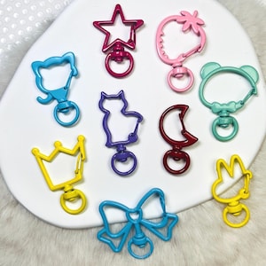 May include: A collection of colorful metal swivel clasps in various shapes, including a star, strawberry, bear, cat, crown, bow, and crescent moon. The clasps are in shades of pink, blue, yellow, and red, and are arranged on a white surface.