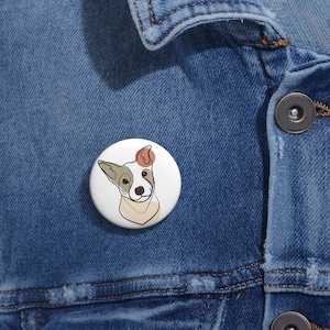 May include: A white button with a line drawing of a dog's head. The dog has brown, white, and orange fur. The dog is looking directly at the viewer.