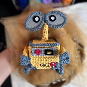 May include: A handmade crochet figure of the character WALL-E from the animated film. The figure is primarily yellow with gray and blue accents. The eyes are large and gray with black and white centers. The name "WALL E" is stitched on the front.