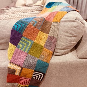 Könnte beinhalten: Eine bunte Patchwork-Strickdecke, die über einem Sofa liegt. Die Decke besteht aus quadratischen und rechteckigen Blöcken in Blau-, Gelb-, Rosa-, Orange- und Brauntönen. Das Sofa hat eine neutrale beige Farbe.