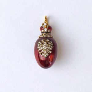 May include: A pendant shaped like an egg with a deep red translucent enamel finish. The piece features a gold-toned double-headed eagle emblem encrusted with clear stones on the front and a decorative crown topper set with small clear stones.