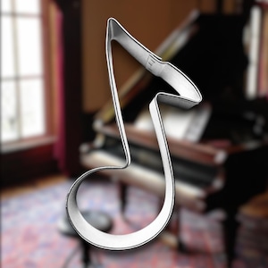 May include: A silver metal cookie cutter shaped like a treble clef. The cookie cutter is in focus, while a blurry grand piano is in the background.