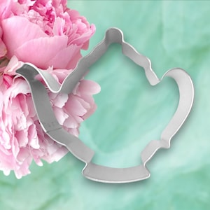 May include: A silver metal cookie cutter shaped like a teapot. The cutter is on a light blue background with pink flowers.