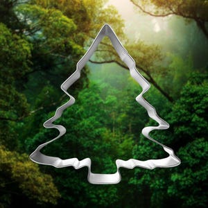 May include: A silver metal cookie cutter shaped like a Christmas tree. The cutter is in focus against a blurry background of green trees.