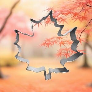 May include: A silver metal maple leaf cookie cutter. The cutter is in focus against a blurred background of red and orange fall foliage.