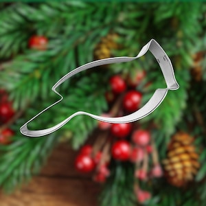 May include: A silver metal cookie cutter shaped like a cardinal bird. The cutter is on a blurry background of green pine branches and red berries.