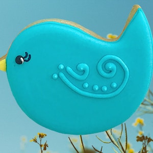 May include: A blue bird-shaped cookie decorated with a white swirl and blue dots. The cookie is on a background of green and yellow plants.
