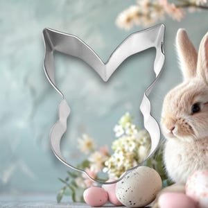 May include: A silver metal cookie cutter shaped like a bunny rabbit head. The cutter is in front of a white bunny rabbit with pink ears and a white and pink speckled Easter egg.