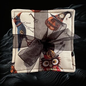 May include: Square fabric coaster with a whimsical Halloween theme. The coaster features a patchwork design with illustrations of a blue bird, an owl, keys, and a witch's hat. A black sheer ribbon is tied around the coaster.