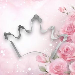 May include: A silver metal cookie cutter shaped like a crown. The cutter is on a pink background with a floral design.