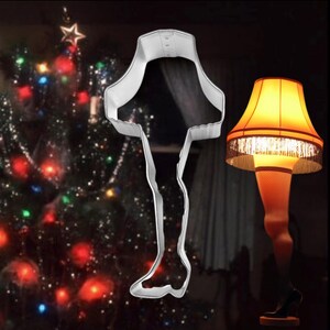 May include: A metal leg-shaped cookie cutter next to a lamp shaped like a leg in a fishnet stocking. The lamp has a yellow lampshade with fringe. A Christmas tree with colorful lights is in the background.