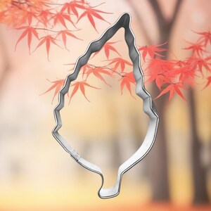 May include: A silver-colored leaf-shaped cookie cutter. The cutter is in front of a blurred background of red maple leaves and a soft, warm-toned backdrop. The image evokes an autumn theme.