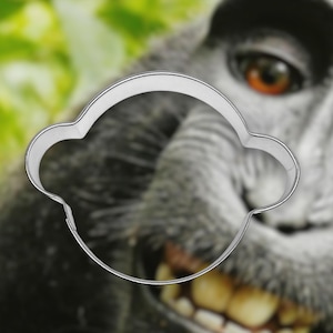 May include: A silver-colored metal cookie cutter in the shape of a monkey's head. The cutter is in front of a blurred image of a monkey's face with a green background. The monkey's face is gray and has a wide grin.