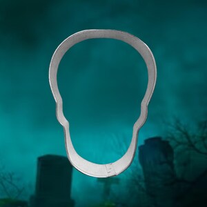 May include: A silver metal skull cookie cutter. The cutter is shaped like a skull with a rounded top and a pointed chin. The cutter is isolated against a blurry background of a graveyard.