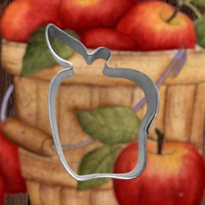 May include: A silver metal apple-shaped cookie cutter. The cookie cutter is in focus and is in front of a basket of red apples.