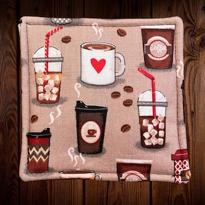 May include: Square beige fabric coaster with coffee-themed illustrations. Features images of coffee cups, iced coffee, coffee beans, and a mug with a red heart. The coaster has a stitched border and is set against a wooden background.
