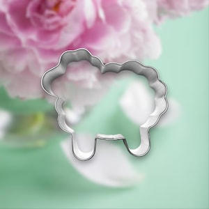 May include: A silver-colored sheep-shaped cookie cutter. The cutter has a wavy outline for the sheep's wool and a simple design for the legs. The background features blurred pink flowers and a light green surface.