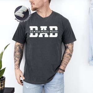 May include: Dark gray t-shirt with the word "DAD" in large white block letters. Below the word "DAD" are the names Josh, Mike, Charlie, and Natalie. The shirt is a casual style.
