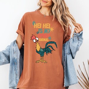 A woman wearing a t-shirt with a cartoon rooster graphic and text, standing in front of a plant.