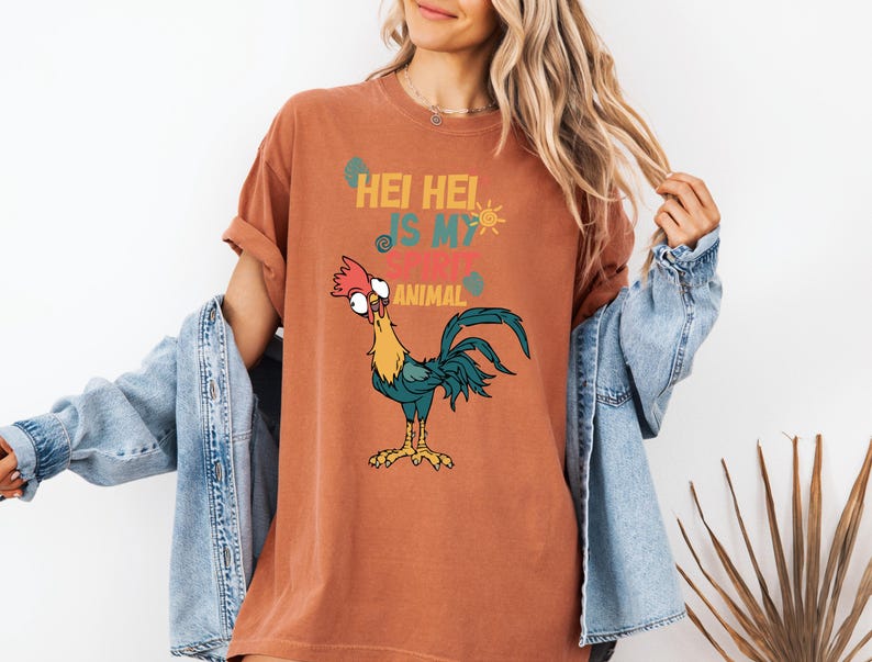 A woman wearing a t-shirt with a cartoon rooster graphic and text, standing in front of a plant.