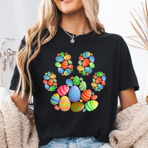 Op de afbeelding: Zwart T-shirt met een pootafdrukontwerp gemaakt van kleurrijke paaseieren. De eieren zijn versierd met patronen en kleuren, waaronder rood, blauw, groen en geel. Een vrolijk ontwerp voor de paasvakantie.