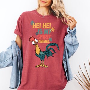 A woman wearing a red t-shirt with a cartoon rooster and text, standing in front of a plant.