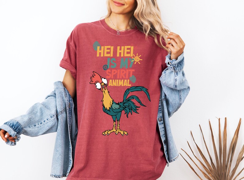 A woman wearing a red t-shirt with a cartoon rooster and text, standing in front of a plant.
