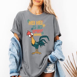 A woman wearing a gray t-shirt with a graphic of a rooster and text, along with denim clothing and a denim jacket.